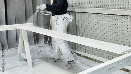 Worker spraying white paint on wooden board in industrial workshop.
