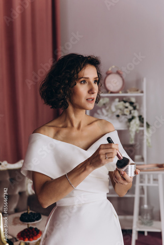 Bride in an off-shoulder gown applies face powder with a brush while getting ready in a softly lit, stylish room. Bride Applying Makeup in Elegant Bridal Suite