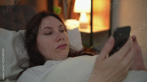 Young adult Caucasian woman lying in bed at night and watching social media feed on smartphone with crooked neck before going to sleep. Concept of digital gadgets and internet addiction