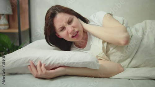  Young adult woman wakes up in the morning in bed and suffers from neck pain. Concept of problems with the spine due to improperly fitted pillow