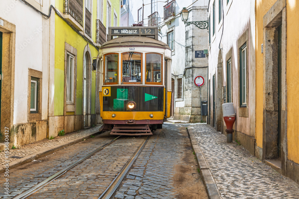 Fototapeta premium A classic yellow Lisbon tram (Elétrico 28) on a charming cobblestone street, surrounded by colorful historic buildings. Quintessential European charm.