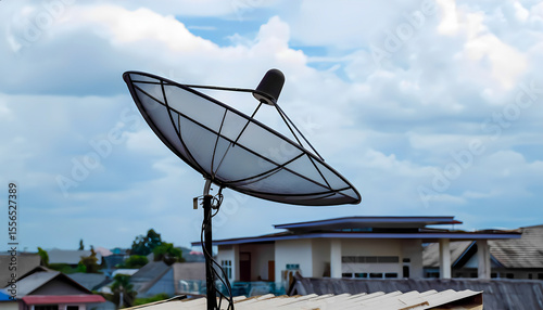 black satellite dish mounted on the roof of a building. The antenna is pointed upward, against a clear blue sky with fluffy white clouds. The design of the antenna includes a mesh dish and an elongate