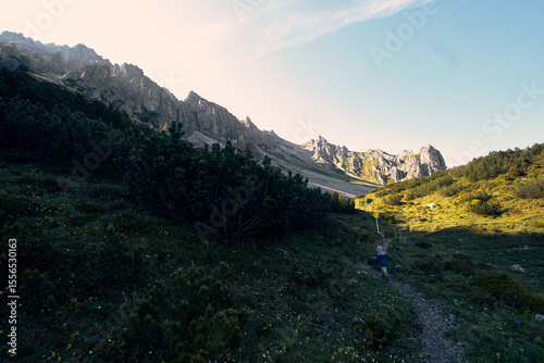 Hiker explores stunning alpine landscape with towering peaks and lush greenery during early evening hours