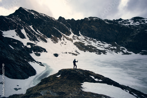 Hiker exploring snowy alpine landscape near frozen lake with dramatic mountain backdrop in the Alps during early spring