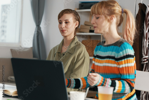 Two young adult women colleagues working together on a laptop in a modern workplace, socializing and collaborating in a friendly business environment, Gen Z professionals.