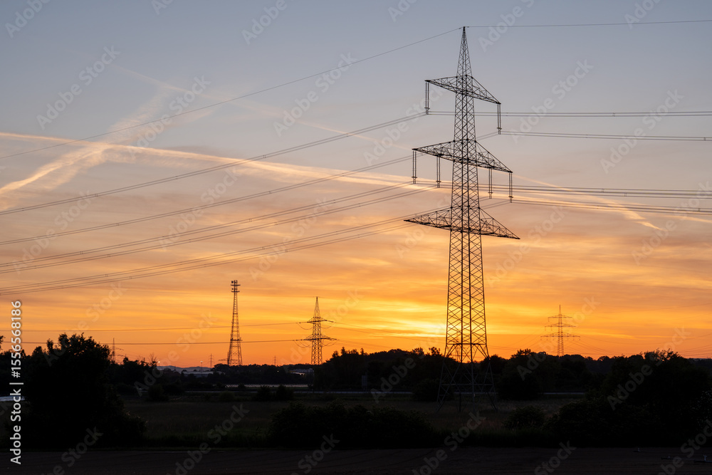 An electricity pylon stands tall as power lines stretch across a vibrant evening sky, with pink and orange hues illuminating layered clouds above a leafy horizon An electricity pylon stands tall as power lines stretch across a vibrant evening sky, with pink and orange hues illuminating layered clouds above a leafy horizon