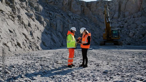 Two engineers in safety vests and hardhats collaborate, analyzing data on a digital tablet within a quarry, with an excavator in the background