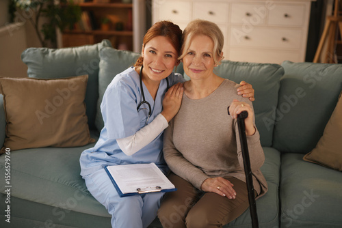 A home healthcare nurse in scrubs smiles while embracing a senior woman who is holding a cane. They are sitting on a sofa in the woman's living room during a daytime visit.