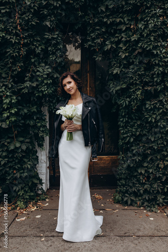 Full-length portrait of a bride in a white gown and leather jacket holding flowers while standing near a lush ivy-covered wall. Bride in Leather Jacket Holding Flowers Near Ivy Wall