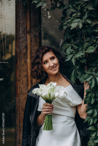 Full-length portrait of a bride in a white gown and leather jacket holding flowers while standing near a lush ivy-covered wall. Bride in Leather Jacket Holding Flowers Near Ivy Wall