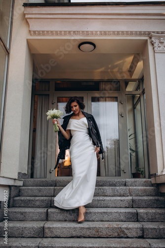 Stylish bride wearing a white wedding dress and a black leather jacket posing confidently on stone stairs in front of a modern building. Modern Bride in Leather Jacket Posing on Urban Steps