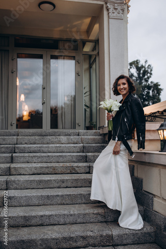 Stylish bride wearing a white wedding dress and a black leather jacket posing confidently on stone stairs in front of a modern building. Modern Bride in Leather Jacket Posing on Urban Steps