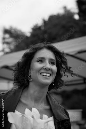 Black and white portrait of a smiling bride wearing a leather jacket and holding flowers, captured in a candid outdoor moment.  Smiling Bride in Leather Jacket — Black and White Portrait
