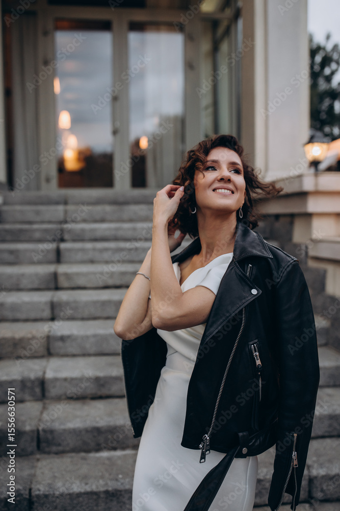 Fototapeta premium Stylish bride wearing a white wedding dress and a black leather jacket posing confidently on stone stairs in front of a modern building. Modern Bride in Leather Jacket Posing on Urban Steps