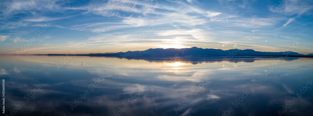 Fototapeta premium Sunset overSalton Sea, Mecca, California, USA. The sun reflects off the calm water, creating a mirror image of the sky and mountains. The scene evokes peace and tranquility.