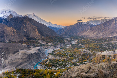 A stunning golden hour view of a mountain valley with a turquoise river, lush autumn foliage, and snow-capped peaks. A breathtaking fusion of nature’s beauty and a serene rural landscape.