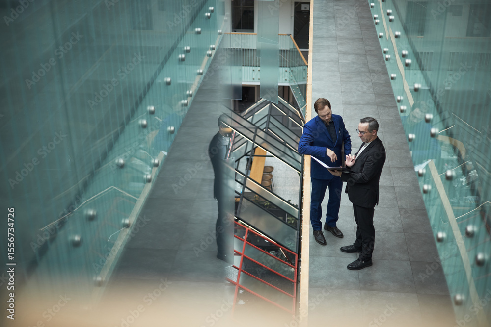 Obraz premium Two businessmen reading and discussing document in a modern office environment with a glass railing and multiple levels, highlighting professional attire and conversational engagement