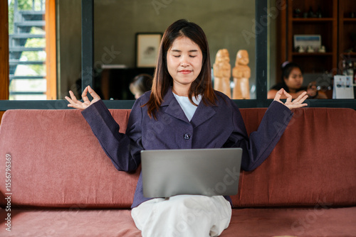 A young woman practices meditation while working on her laptop, promoting relaxation and mindfulness in a modern living room setting.