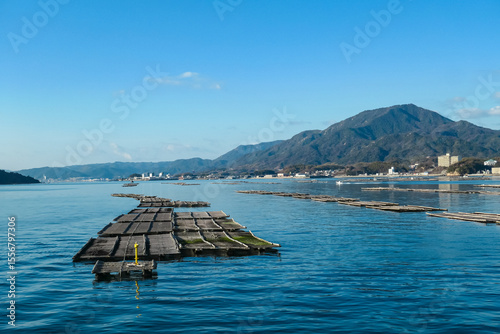Wooden oyster farms float on the serene blue waters of Seto Inland Sea near Miyajima, Japan. This picturesque view shows the traditional aquaculture industry against a backdrop of lush green mountains