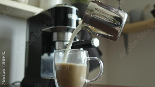 Milk pours from a pinscher into a glass cup of coffee. Cappuccino preparation. There is a coffee machine in the background