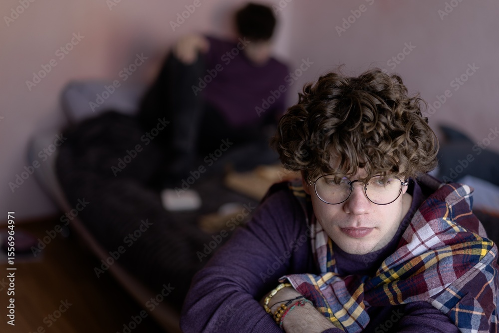 Obraz premium Young man with curly hair gazes thoughtfully while friend relaxes on bed in cozy, softly lit room during quiet afternoon
