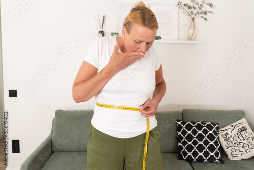 Quadro su tela Surprised mature woman measuring her waistline with tape, expressing shock about sudden weight gain or bloating, in a healthy living context at home