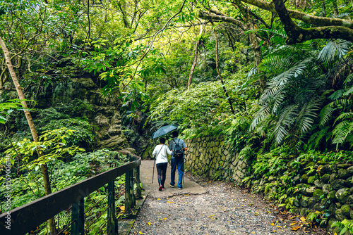 An older couple walks along a metal barrier walkway between mossy rocks. She carries a grey backpack and a beige bucket hat, surrounded by lush green trees in Taroko National Park, Taiwan.