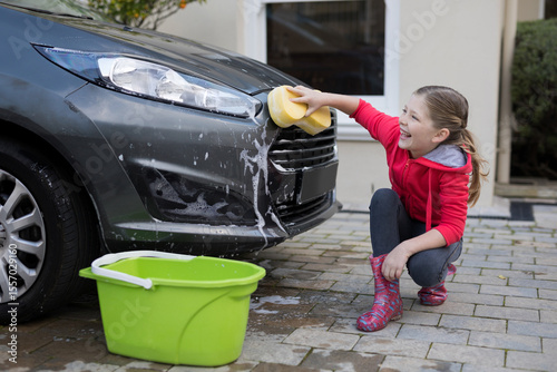 Child in red hoodie scrubbing car bumper with yellow sponge and bucket on driveway, copy space