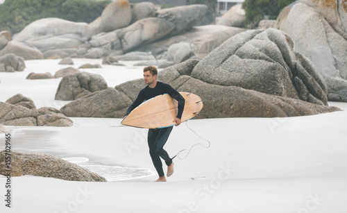 Adult male wearing black wetsuit walking across white sand carrying wooden surfboard with leash