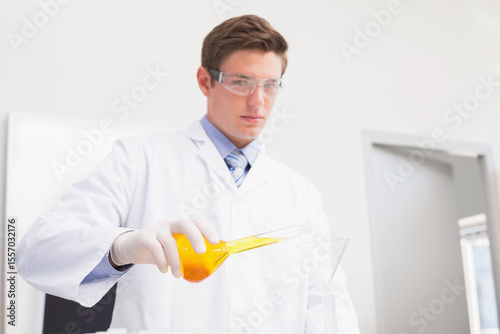 Male scientist in white coat pouring orange liquid through funnel into glass container on lab bench