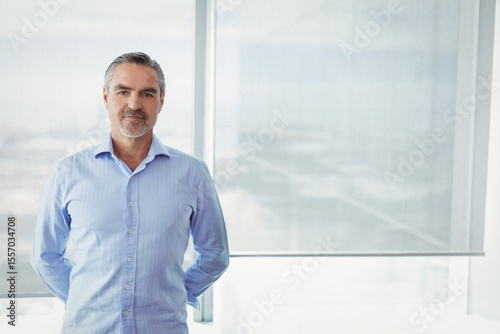 Businessman standing with hands behind back by office window roller blind diffusing cityscape light