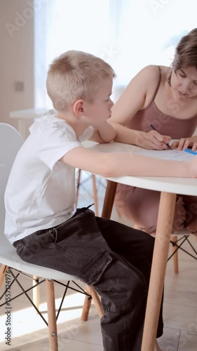 A boy with a hearing aid and his mother sit together in the kitchen or living room, working on homework. A natural, heartwarming scene of bonding, learning, and everyday family life. Authentic emotion