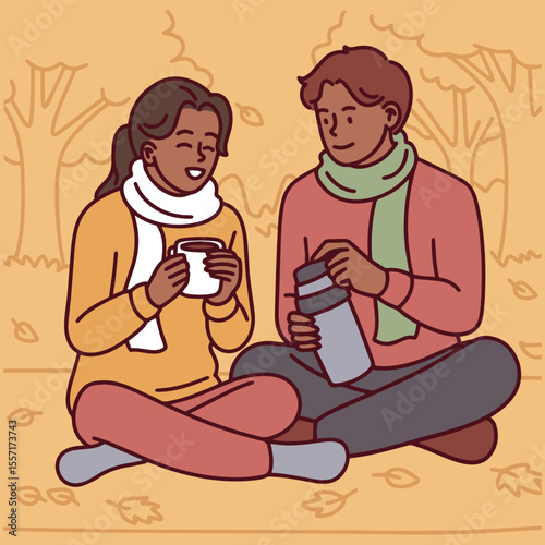 Dark-skinned couple dressed in warm clothes sits on ground in autumn park, drinking hot beverages together from thermos in a calm fall scene