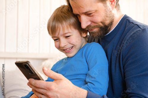 Father and son little boy laughing and using phone. They watching funny videos together, playing games on smartphone. Concept technology, smart phone addicted. Fun weekend activity. Modern fatherhood