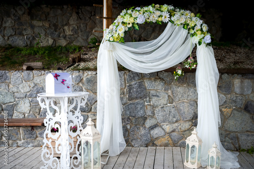 Decorated wedding arch. A decorative wedding arch is adorned with flowers and drapes, set against a stone backdrop and wooden flooring.