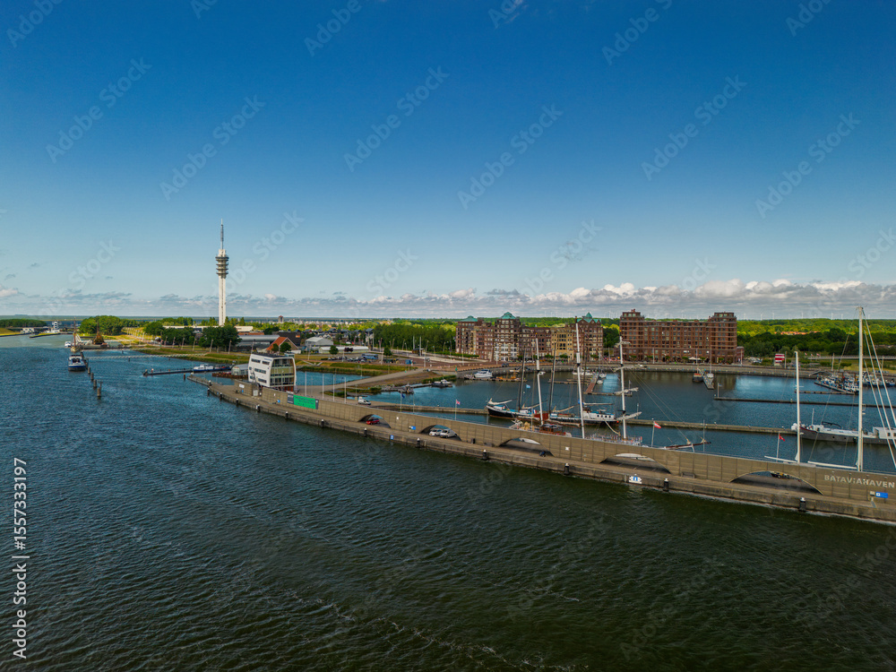 Fototapeta premium A scenic harbor view with sailboats docked along the pier. A tall communication tower stands in the distance. Red-brick residential buildings and lush green trees frame the calm waterfront.