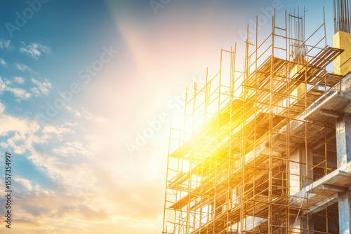 A building under construction with metal scaffolding is illuminated by the warm glow of the setting sun against a blue sky