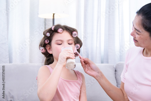 Kids, cosmetic, child, beauty, makeup, A girl with hair curlers drinks dairy from a glass while her mother applies blush, enjoying fun family moments and learning selfcare in relaxed living room