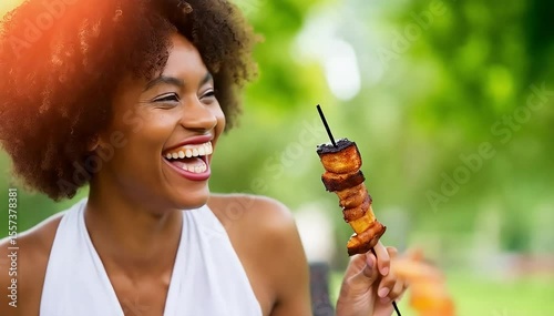 A joyful woman with an afro hairstyle laughs heartily while enjoying a barbecue skewer at a sunny outdoor party