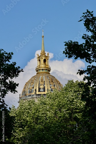 The magnificent golden dome and spire of Les Invalides in Paris shine brightly against a blue sky with clouds, beautifully framed by lush green trees, highlighting its intricate details