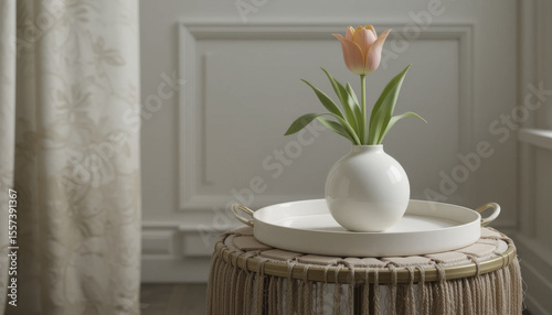 single pink tulip in white vase sits on round tray atop woven stool, creating serene and elegant atmosphere in softly lit room
