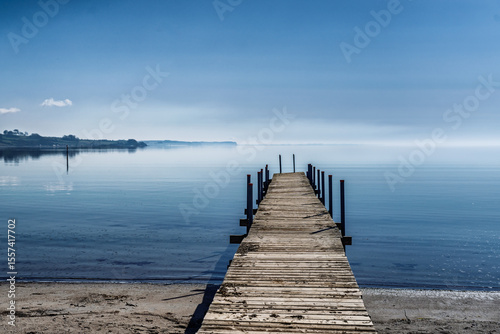 Fototapeta Naklejka Na Ścianę i Meble -  Wooden Pier Extending Over Peaceful Water Under Blue Sky, Vemmingbund, Denmark