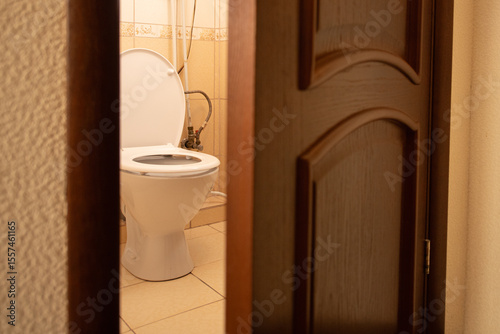 A clean bathroom features a white toilet seen through an open wooden door. The floor is tiled, and the walls have a subtle design, giving a simple yet comforting look
