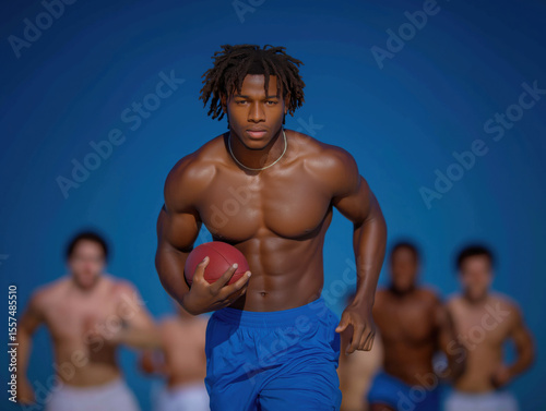 Athletic young man with muscular build running on field while holding a football, surrounded by teammates in background, showcasing teamwork and competitive spirit