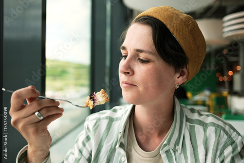 A young pretty woman examines a bite of cake on a fork while sitting by a bright window in a modern cafe.