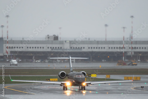 Business jet taxiing on wet runway in rainy weather, front view of private executive aircraft with landing lights on and blurred airport terminal in background