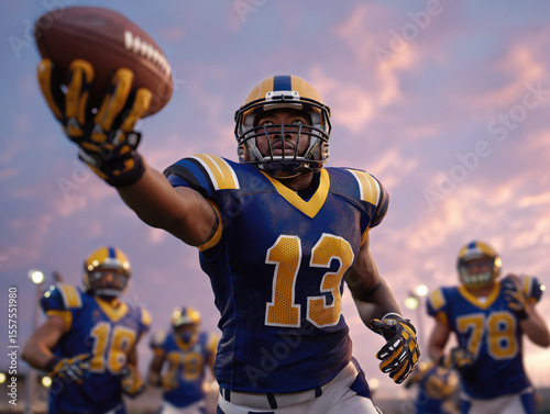 Male football player in blue and yellow uniform reaching out to catch a football during an evening practice session with teammates in the background showcasing teamwork and athleticism