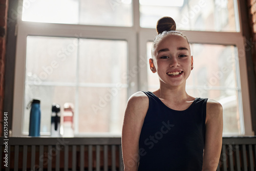 Portrait of Caucasian girl smiling at camera during rhythmic gymnastics training session in studio, hair in bun, wearing leotard, natural light from large window behind Free Download Portrait of Caucasian girl smiling at camera during rhythmic gymnastics training session in studio, hair in bun, wearing leotard, natural light from large window behind - GFXLoad