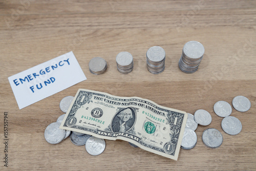 Clear jar marked emergency fund with stacked coins, folded dollar bills, and loose change on wooden table.