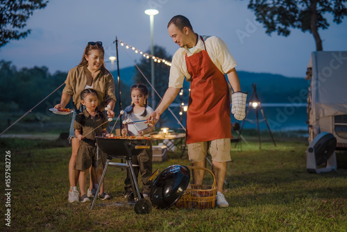 A man is cooking food on a grill with a woman and two children. The man is wearing an apron